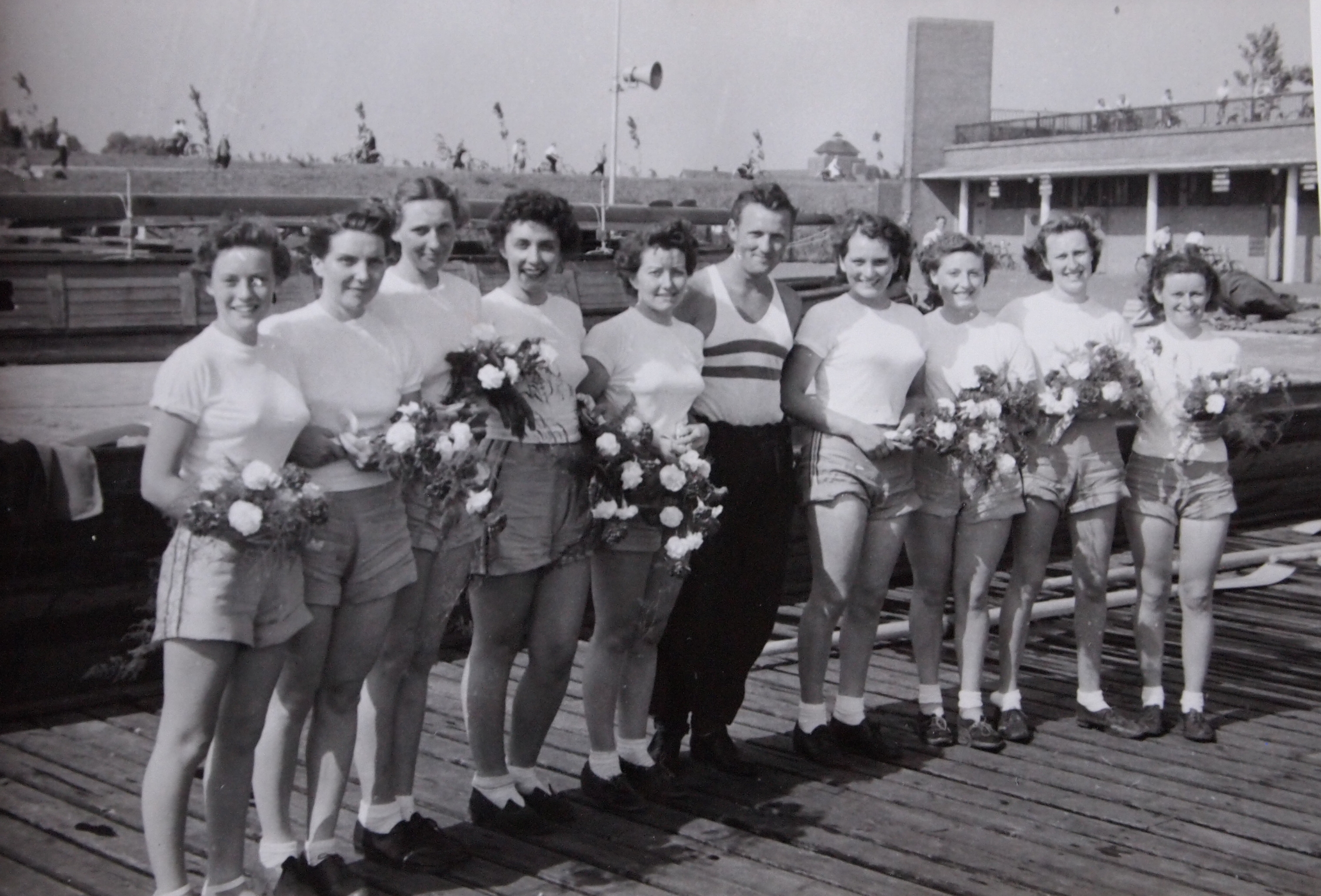 b/w photo sof women's eight holding bunches of flowers on landig stage with male coach
