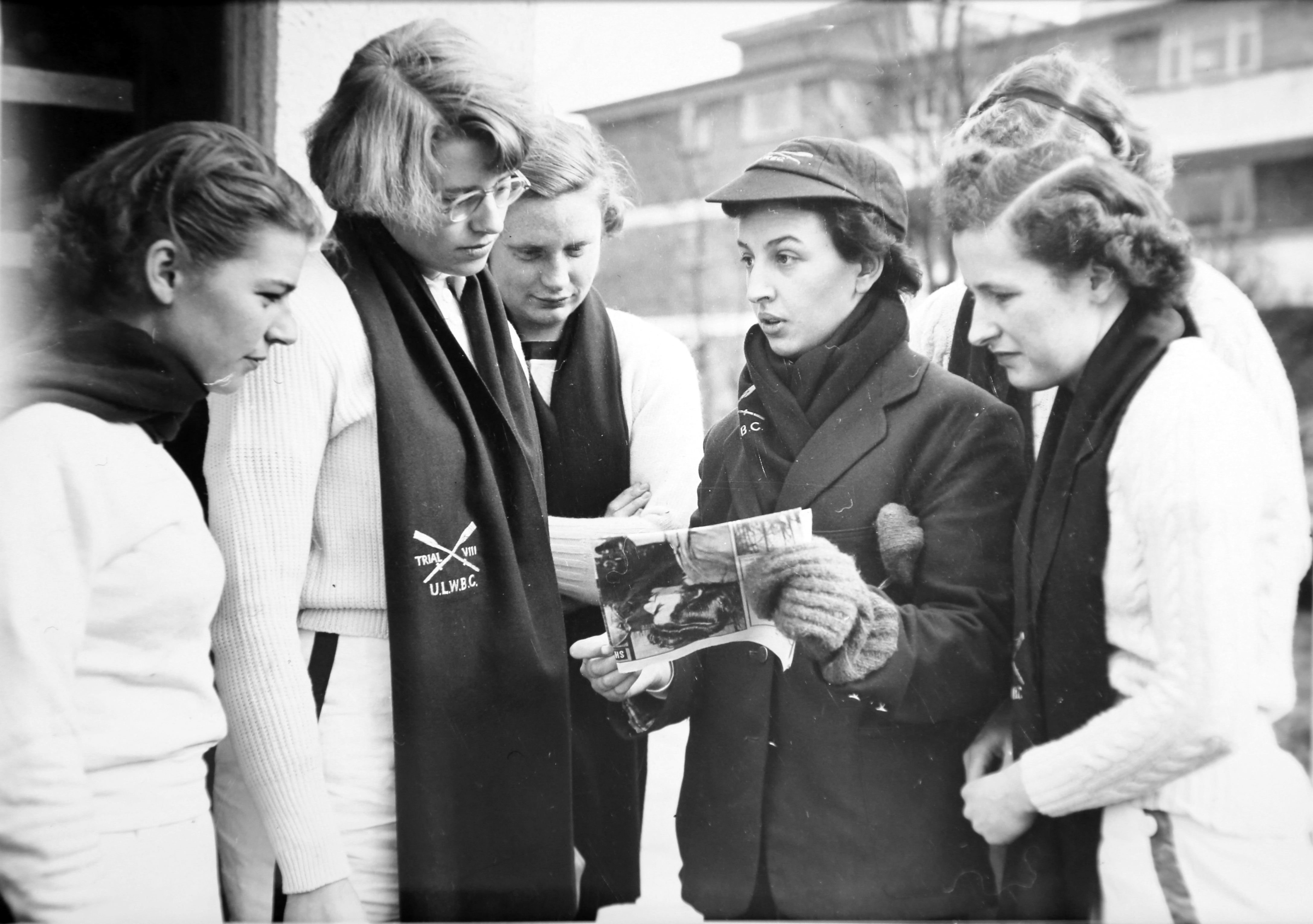 5 rowing women and cox looking at newspaper clipping