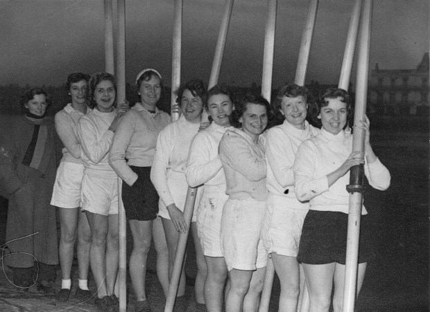 Eight women in shorts and white tops with oars vertical, cox in long coat and scarf