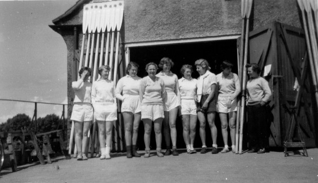 9 women with oars leaning up against boathouse