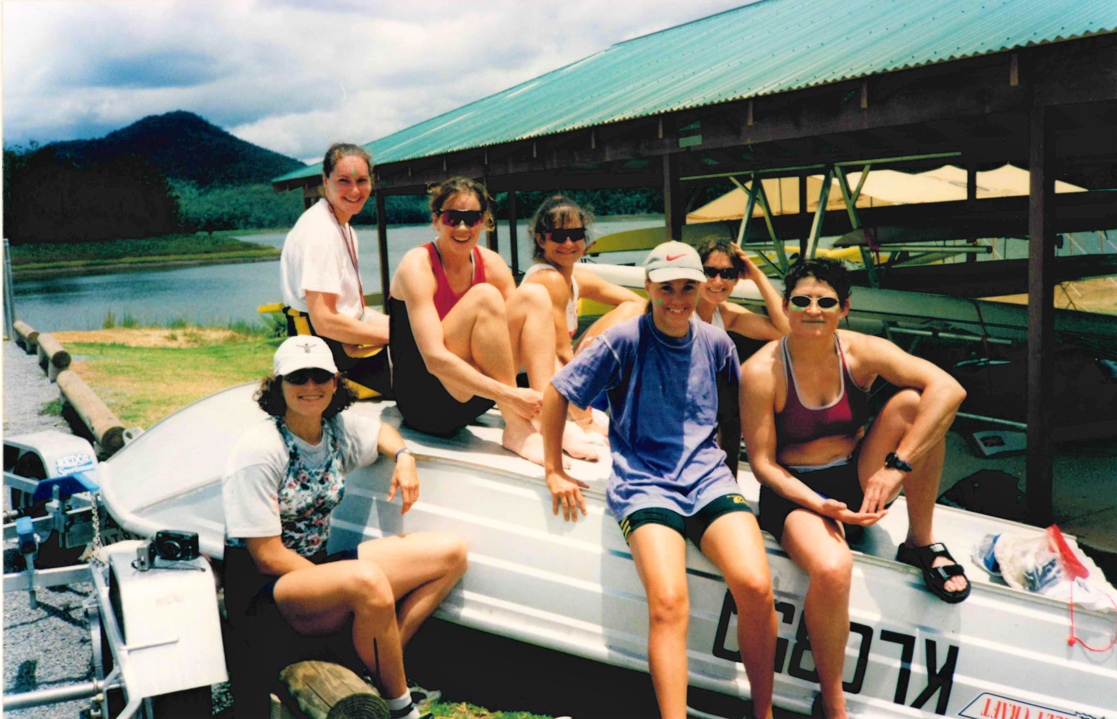 7 women sitting on upturned coaching launch by boat racks
