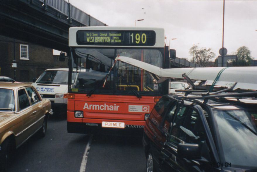 Red bus with sculling boat on roof of car in front bent sideways on windscreen