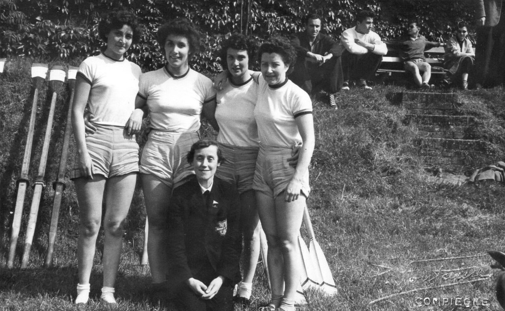 Black & white photo of 5 women - 4 rowers in white shorts and cox in dark blazer.