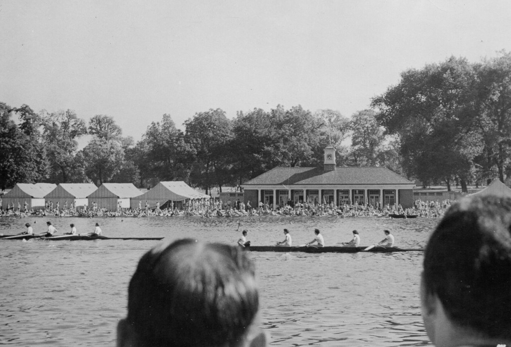 Women's fours racing on the Serpentine with pavillion in the background and men's heads in the foreground
